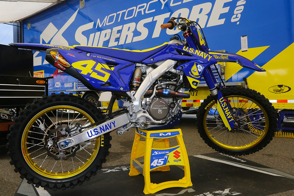 Motocross racing bike with blue and gold graphics against a backdrop of Motocross Super Store banner