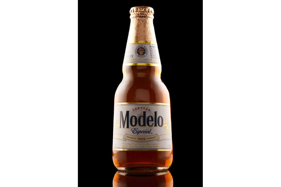 棕色玻璃啤酒瓶，白色标签上印有 Modela，瓶口附近靠近瓶盖的地方还有附加标签